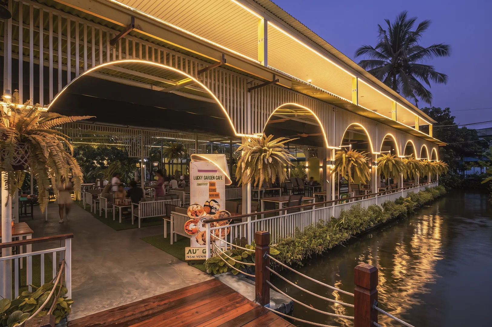 Lucky Garden Cafe pavilion lit up at dusk with arched LED lights alongside a canal lined with tropical plants.