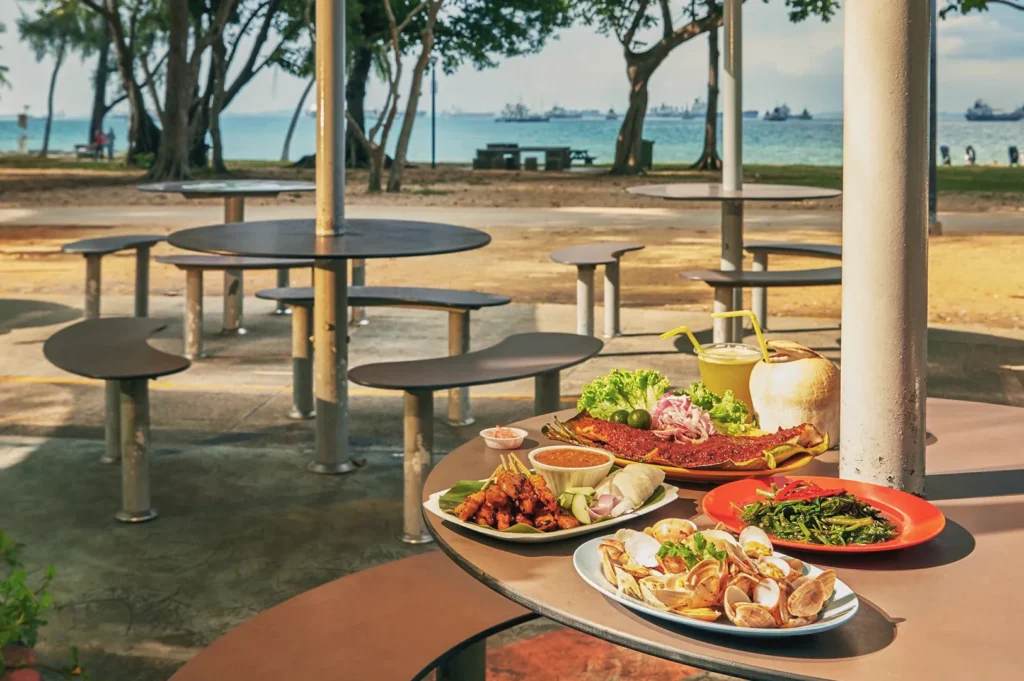 Outdoor seaside dining setup at East Coast Park, Singapore — plates of grilled seafood, satay, and coconut drinks overlooking the beach and ships.