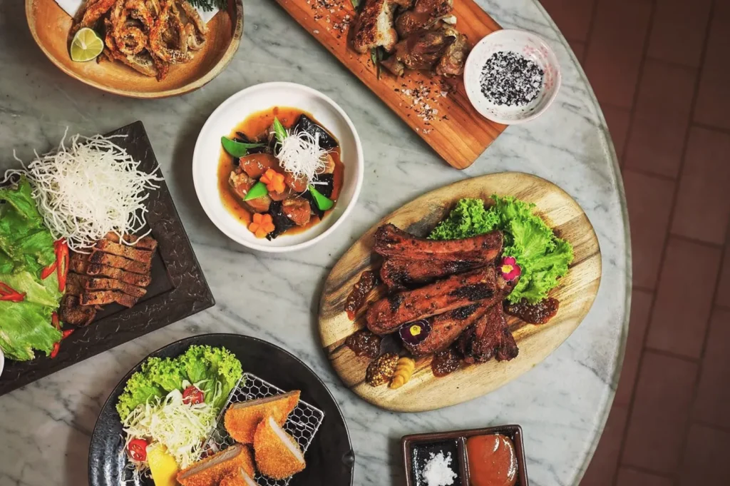 Assorted Asian dishes on marble table including grilled ribs, fried cutlets, and stir‑fried vegetables — gourmet food photography showcasing Singaporean fusion cuisine.