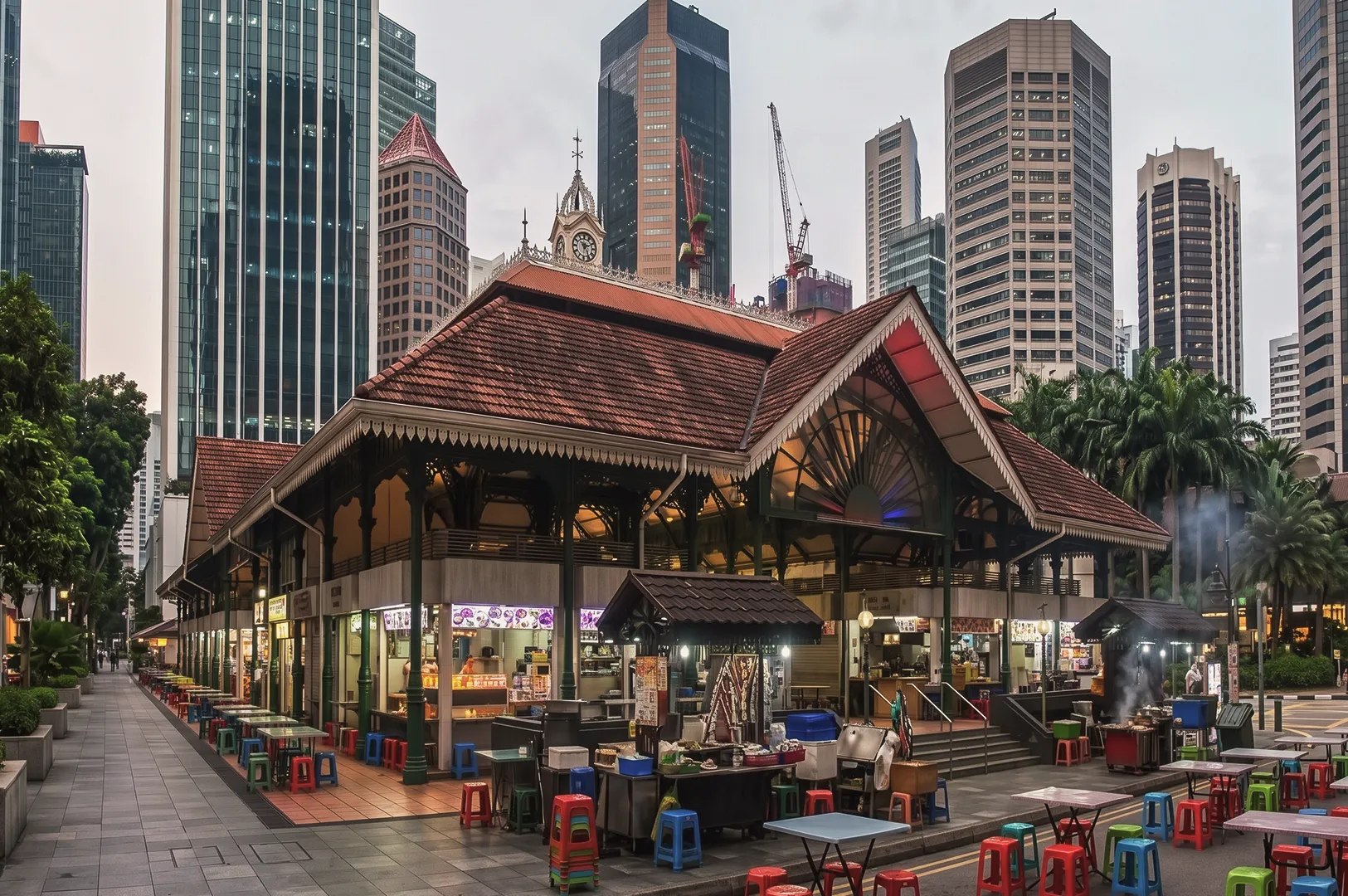 Lau Pa Sat hawker centre in Singapore with red‑tiled roof and ornate colonial architecture surrounded by modern skyscrapers and outdoor dining tables.