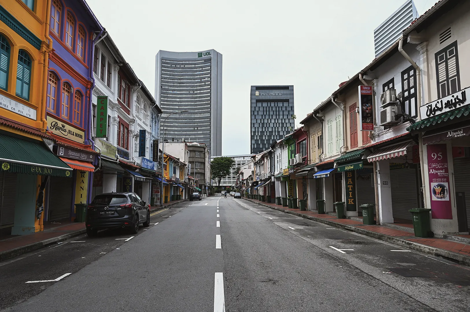 Quiet street framed by colorful historic shophouses in Singapore leading toward modern skyscrapers under an overcast sky.