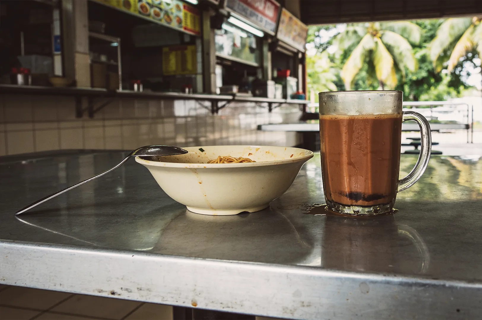 Iced milk tea (teh tarik) in a glass mug alongside a bowl of local noodle soup on a stainless steel table at an open-air Singapore hawker centre.