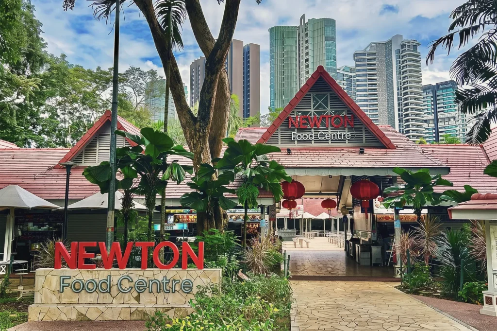 Newton Food Centre Singapore entrance with red roof, tropical plants, and red lanterns surrounded by modern city skyscrapers.