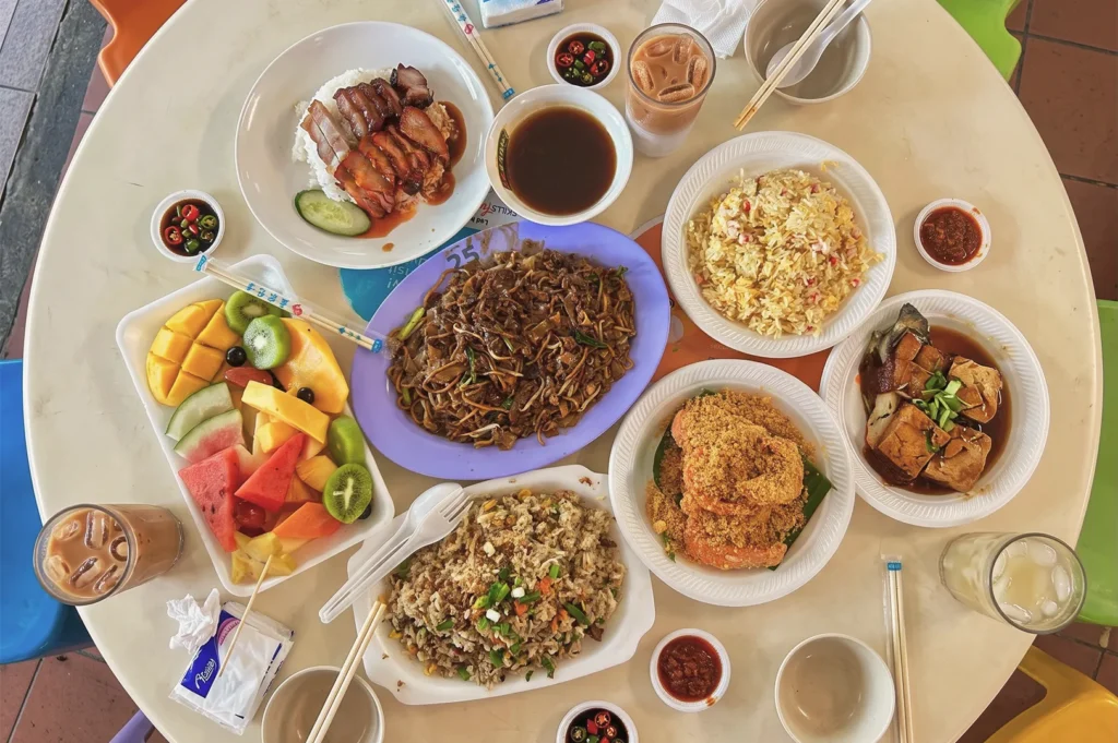Colorful spread of Singapore hawker dishes including roasted meats, fried rice, noodles, cereal prawns, tofu, and fresh fruit platter.