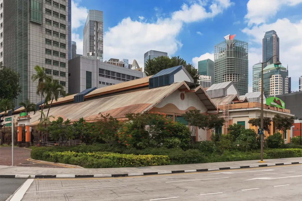 Exterior of Maxwell Food Centre Singapore with heritage façade, tropical greenery, and modern high‑rise buildings in the background under a bright blue sky.