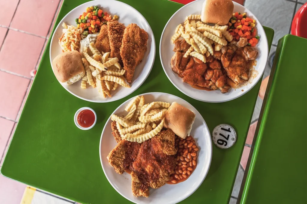 Close‑up of three plates of Western food at Maxwell Food Centre Singapore featuring fried chicken, fish fillet, crinkle fries, baked beans, and mixed vegetables.