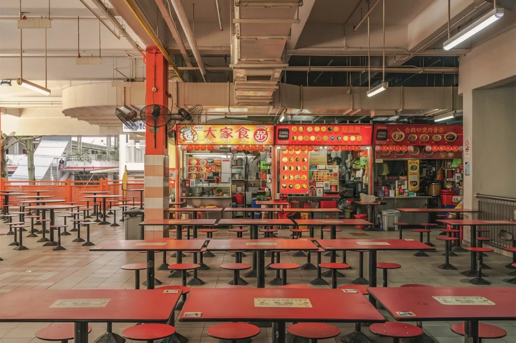 Empty Singapore hawker centre dining area filled with red tables and matching round stools in front of illuminated food stalls.   