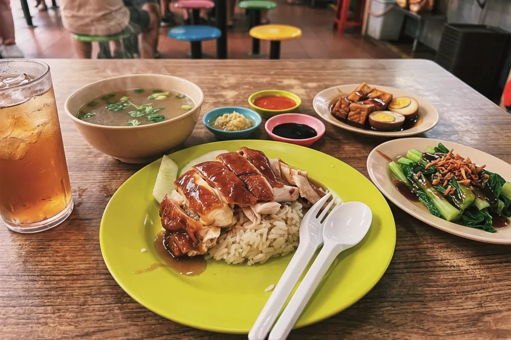 Singapore hawker meal featuring roasted chicken rice with soup, iced tea, braised tofu, and stir‑fried vegetables served on colorful plates.