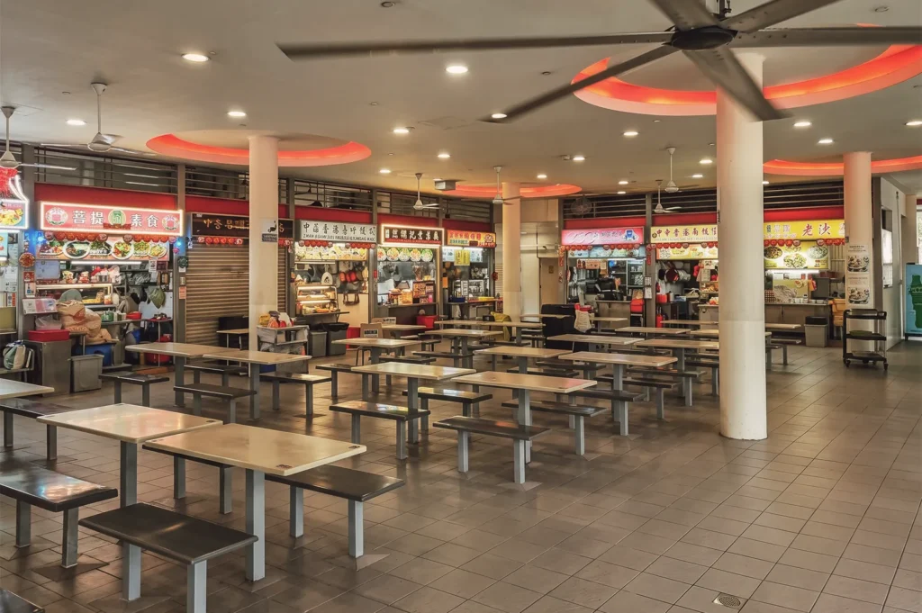 Singapore hawker centre interior with rows of tables, bright food stall signs in Chinese, and ceiling fans under warm lighting.