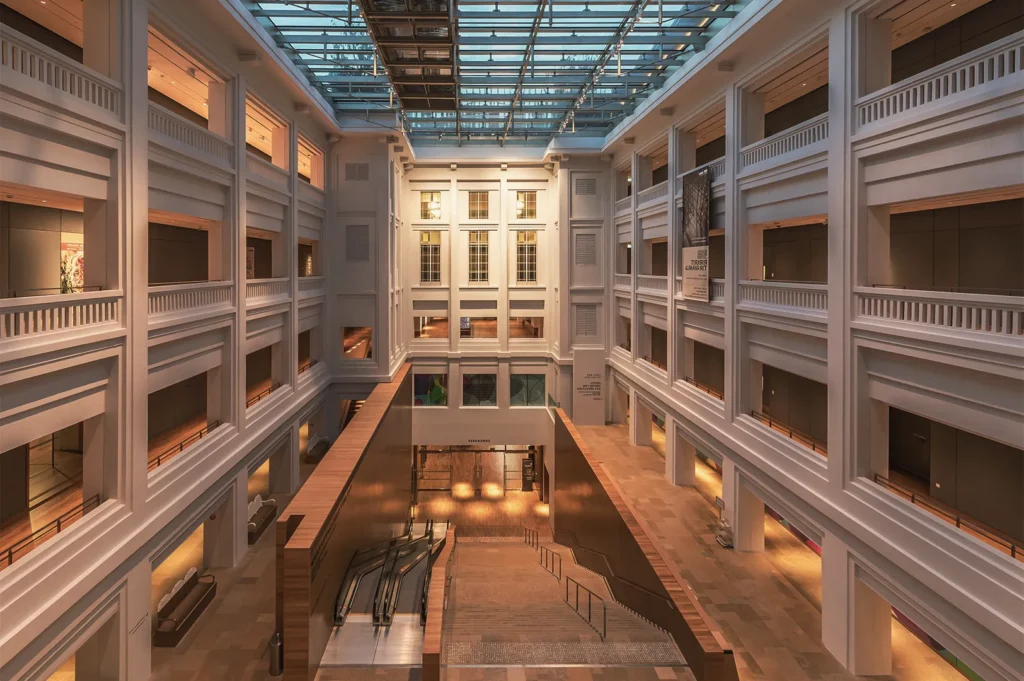 Expansive glass‑roofed atrium with symmetrical balconies, escalators, and warm lighting blending classical and contemporary design elements.