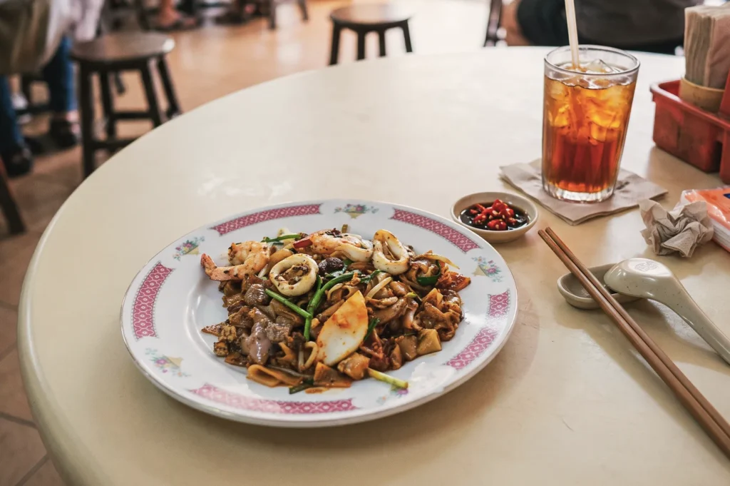 Plate of Singapore char kway teow noodles with prawns, squid, and fish cake served with iced tea and chili sauce in a hawker setting.