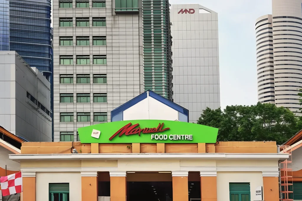 Front entrance building of Maxwell Food Centre featuring a bright green signage board set against modern high-rise city skyscrapers in Singapore.