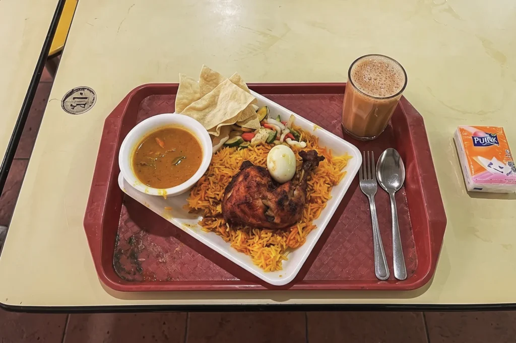 Singapore hawker meal of chicken biryani with curry, papad, boiled egg, and teh tarik served on a red tray.